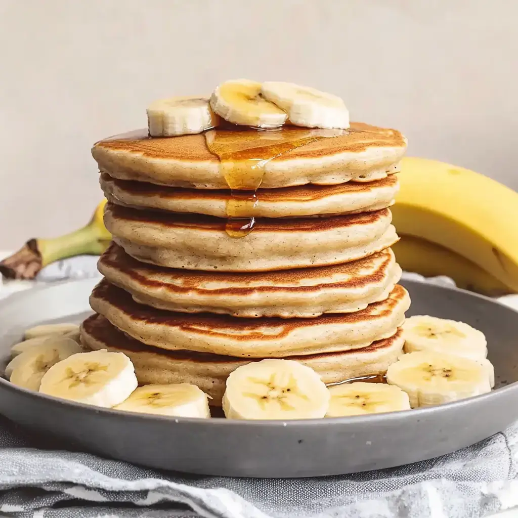 Auf einem Teller liegt ein dicker Turm Pfannkuchen mit Banane und Sirup. Daneben wartet eine frische Banane.