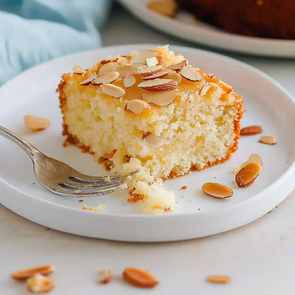 Auf einem weißen Teller liegt ein Stück saftiger Mandelkuchen, bestreut mit Mandelblättchen, daneben eine Gabel.