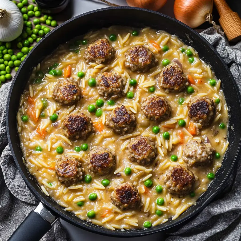 In der Pfanne liegen Frikadellen, kleine Nudeln, Erbsen und Karotten in cremiger Sauce.