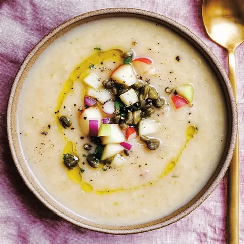 Cremige Suppe in einer Schale, garniert mit Apfelwürfeln, Kapern und Olivenöl, auf rosa Stoff.