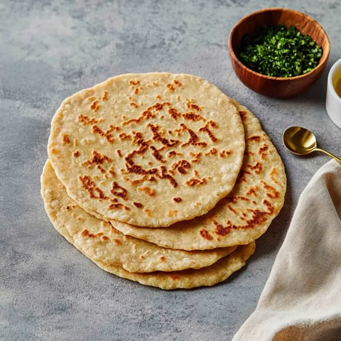 Auf einer grau-marmorierten Platte stapeln sich frisch gebackene Fladen; daneben steht ein Schälchen mit frischen Kräutern.