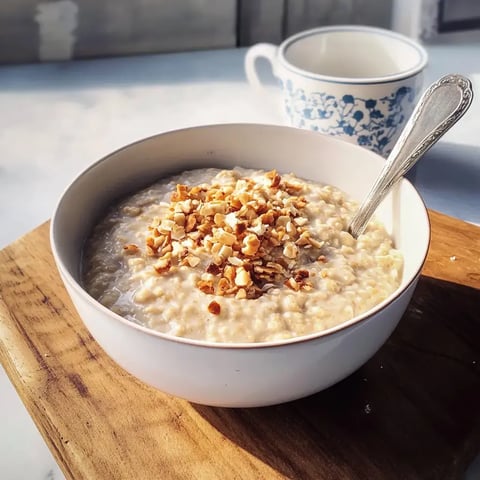 Schüssel Haferbrei mit Nüssen oben drauf steht auf einem Brett neben einer Kaffeetasse.