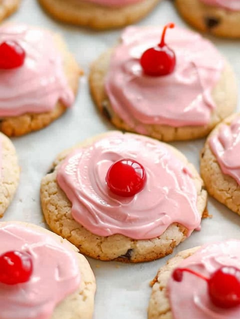 A plate of cookies with pink frosting and cherries on top.